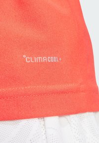 Pomarańczowa koszulka sportowa z napisem "CLIMACOOL+" w kolorze białym na dolnym brzegu. Materiał wydaje się lekki i ma teksturowaną powierzchnię.