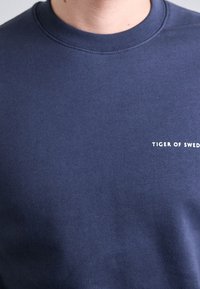 Felpa blu navy in cotone, con collo a girocollo ribattuto e testo del logo "TIGER OF SWEDEN" in bianco sul fronte. Tessuto liscio.