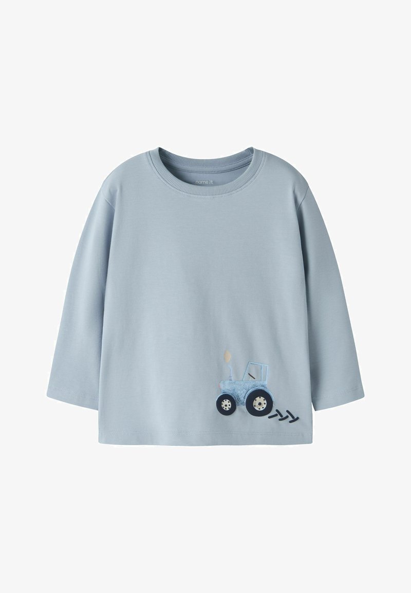 Φωτεινό γαλάζιο παιδικό πουκάμισο με μακριά μανίκια, με ένα μικρό μπλε τρακτέρ και τρία μαύρα σημάδια από ελαστικό κοντά στο κάτω μέρος.