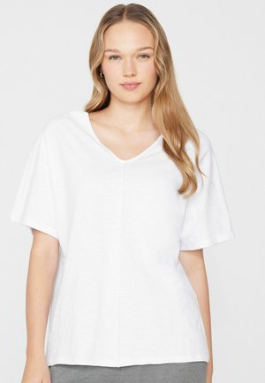 Joven con cabello largo y rubio que lleva una camiseta blanca de manga corta y cuello en V, suelta, sobre un fondo claro y liso.