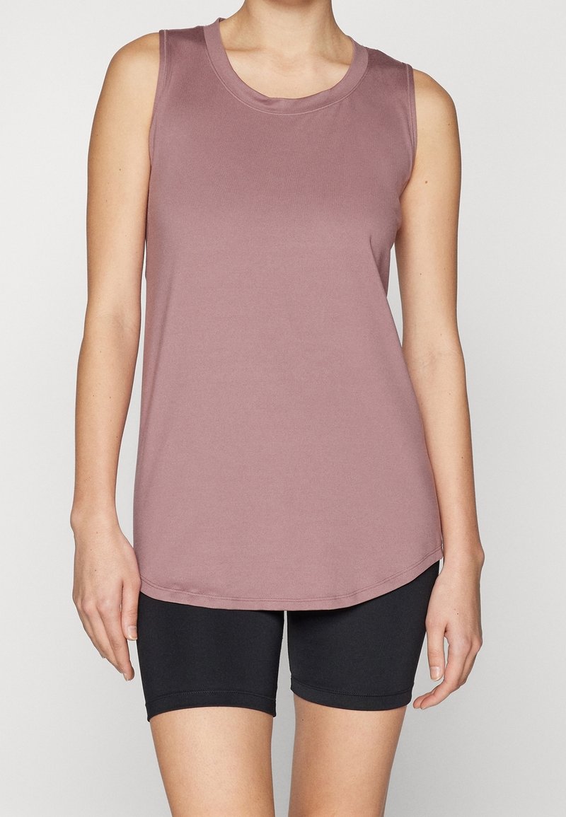 Personne portant un débardeur mauve sans manches et un short noir moulant sur un fond uni, bras détendus le long du corps.