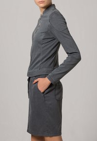 Chaqueta gris con cremallera frontal, mangas largas y acabado texturizado, combinada con una falda a juego que incluye bolsillos y cintura ajustable con cordón.