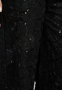 Tessuto nero con motivi floreali in paillettes; texture intricata e brillantezza visibile. Il primo piano mostra i dettagli e le cuciture.