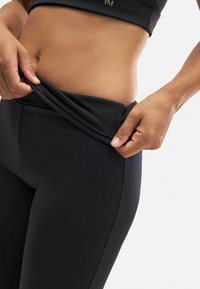Leggings negros de tela elástica, que se muestran siendo tirados hacia abajo ligeramente. Tienen una textura suave y un diseño ajustado.