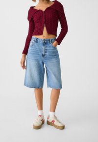 Bordeaux kabelgebreide cropped cardigan met off-shoulder ontwerp, gecombineerd met lichtblauwe denim Bermuda-short en beige sneakers met rode accenten.