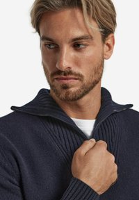 Marine-blaues Zip-Pullover mit geripptem Kragen, gestricktem Textil und kontrastierendem Reißverschluss; Modell hält den Kragen nahe am Hals.