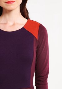 Fioletowa i bordo bluzka z długimi rękawami oraz czerwonym akcentem na ramieniu. Gładka tekstura, dopasowany krój podkreślający linię dekoltu.