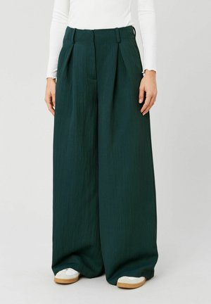 Femme portant un pantalon plissé à jambes larges vert foncé et des baskets blanches, associé à un haut blanc ajusté à manches longues.