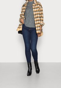 Ruudullinen keltavalkoinen takki harmaan kaapelineulospuseron päällä, yhdistetty tummansinisiin kapeisiin farkkuihin ja mustiin nilkkureihin.