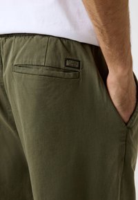 Olivgrüne Hose mit glatter Textur, ausgestattet mit einer Gesäßtasche und einem schwarzen Logoaufnäher. Das Material wirkt leicht und lässig.