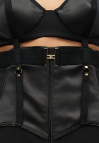 Μαύρο δομημένο μπούστο με ρυθμιζόμενους ιμάντες, κεντρικό φερμουάρ, φαρδιά ελαστική ζώνη με χρυσό κούμπωμα και διακριτικό ανάγλυφο λογότυπο.