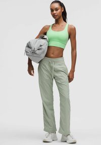 Jasnozielony stanik sportowy oraz blade zielone dresy. Modelka trzyma szary plecak w zaokrąglonym kształcie z detalami zamka błyskawicznego. Białe sneakersy.