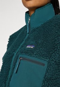 Pullover in pile color teal con collo a zip, superficie testurizzata e tasca sul petto con zip. Presenta il logo Patagonia in alto a sinistra.