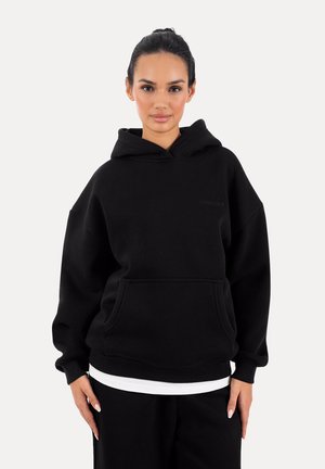 Kobieta o ciemnych włosach związanych do tyłu, ubrana w luźną czarną bluzę z kapturem na białej koszulce, stojąca na tle jednolitego białego tła.