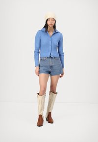 Cardigan côtelé bleu clair, shorts en denim coupés, bottes hautes crème avec des accents marron. Le modèle porte un bonnet en tricot crème. Fond simple.
