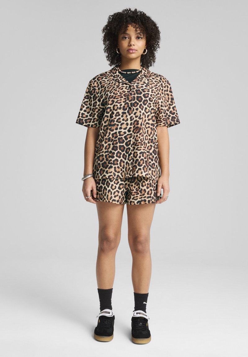 Conjunto de camisa de manga corta con estampado de leopardo y pantalones cortos en tonos beige y negro. Presenta un corte holgado, textura suave y un diseño de botón en la parte delantera.