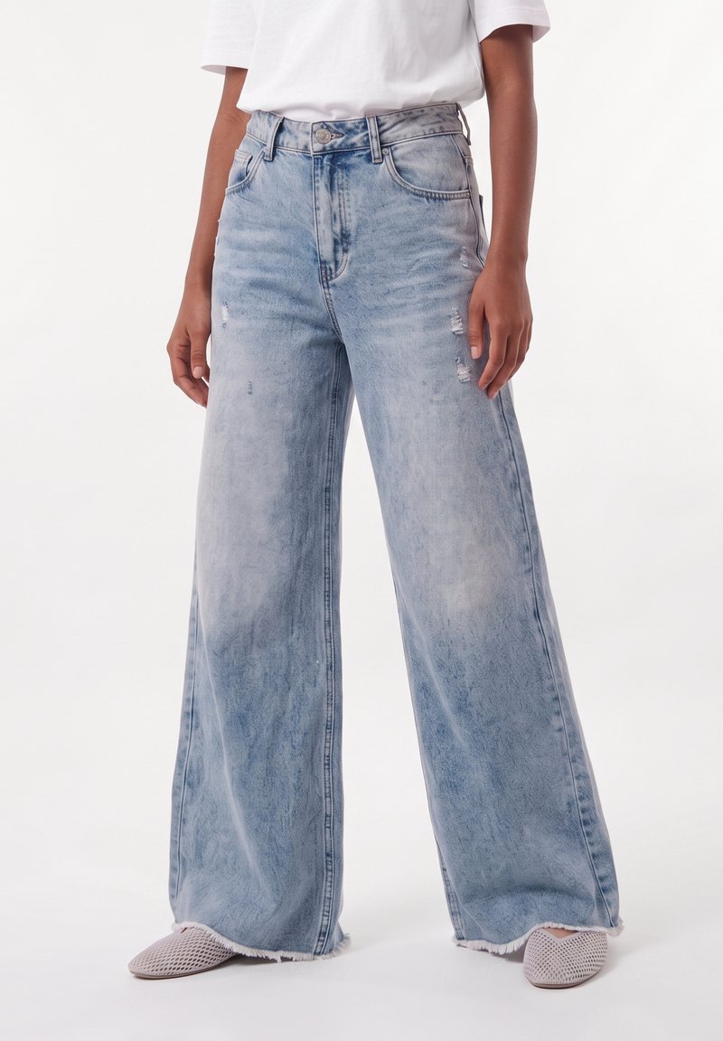 Personne portant un jean bleu clair à jambes larges avec une légère dégradation et des ourlets effilochés, associé à des chaussures beige tissées sans lacets.