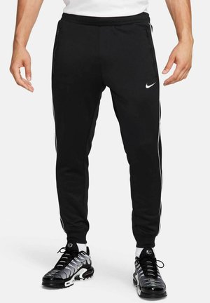Czarne spodnie dresowe Nike z białymi paskami po bokach, kieszeniami na zamek i białym logo Nike na lewej udo, noszone z czarno-białymi butami sportowymi.