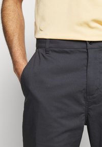 Man wearing dark gray trousers and a light yellow shirt with left hand casually in pocket against plain background.