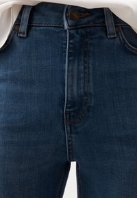 Närbild av midjan på mörkblå denimjeans med knapp, bältesöglor och synliga sömmar under ett instoppat vitt tyg.