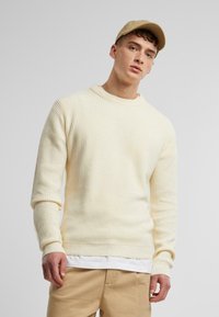 Suéter de punto acanalado color amarillo claro con cuello redondo y mangas largas, combinado con una camiseta blanca y pantalones beige.