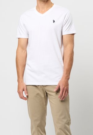 Biała koszulka z dekoltem w serek i krótkim rękawem, z małym czarnym logo po lewej stronie klatki piersiowej. Miękka, lekka bawełniana tkanina.