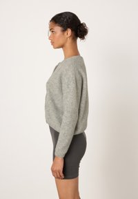 Žena stojící v profilu, oblečená v šedém huňatém svetru a tmavých přiléhavých kraťasech, na jednoduchém světlém pozadí.