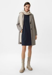 Karierten Mantel mit schwarzen Knöpfen, kombiniert mit einem navyblauen Kleid und einem hellbeigen Rippenschal. Enthält hellbeige klobige Stiefel mit strukturiertem Finish.