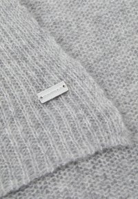 Harmaa neulepuseron yksityiskohta, jonka materiaali on pehmeä ja teksturoitu. Mukana on hopeinen metallilappu, jossa on kaiverrettuna "REPEAT".