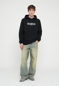 Sudadera negra con bolsillo canguro en la parte frontal y cordón, presenta el logo "DONDUP". Combinada con jeans anchos de lavado claro y zapatos grises.