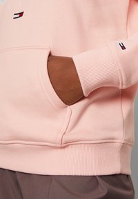 Sudadera de color rosa claro con un bolsillo frontal, puños y dobladillo acanalados; presenta un pequeño acento de logo en colores rojo, blanco y azul.