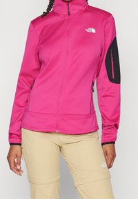 Růžová softshellová bunda s vysokým límcem, zapínacími kapsami a černou nášivkou na rukávu. Lehká tkanina, hladká textura a slim fit design.