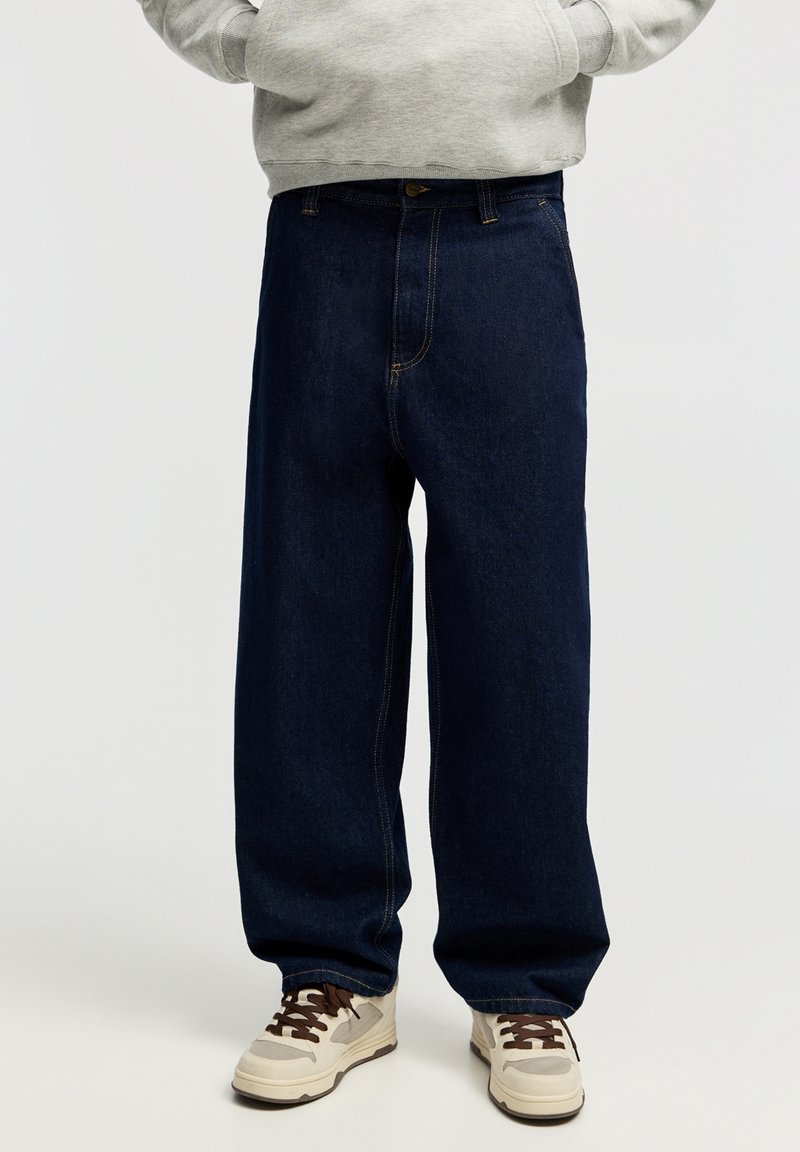 Personne portant un jean large bleu foncé, un sweat à capuche gris clair et des baskets beiges avec des lacets marron, debout sur un fond uni.