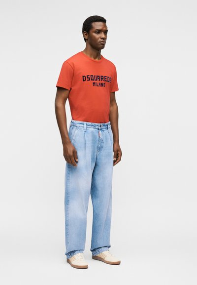 Άνδρας που στέκεται, φοράει πορτοκαλί μπλουζάκι DSQUARED2 Milano, ανοιχτό μπλε άνετα τζιν και λευκά αθλητικά παπούτσια σε μονόχρωμο ανοιχτό φόντο.