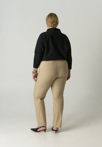 Zwarte blouse met lange mouwen; beige, nauwsluitende broek; zwarte pumps met puntige neus; gladde textuur; minimale hardware; effen witte achtergrond.