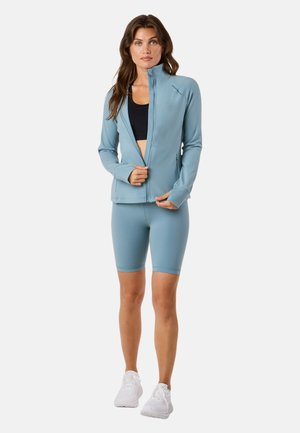 Vrouw met lichtblauwe jas met ritssluiting, bijpassende fietsshort, zwarte sportbeha en witte sneakers, staand tegen een witte achtergrond.