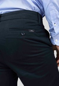 Pantalon ajusté bleu foncé avec une texture lisse, doté d'une poche arrière avec un bouton et d'une étiquette indiquant "Mise Au Green."