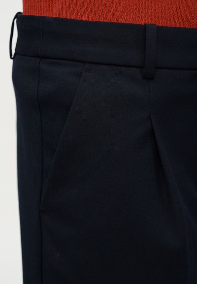 Κοντινή λήψη σκούρων μπλε σκούρων παντελονιών με πιέτες και ζώνη μέσης, που φοριούνται με υφασμάτινο κόκκινο τοπ.