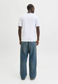 Valge puuvillane T-särk lühikeste varrukatega, ühendatud laia lõikega siniste teksadega. Teksadel on viie taskuga disain ja nähtav õmblemine.