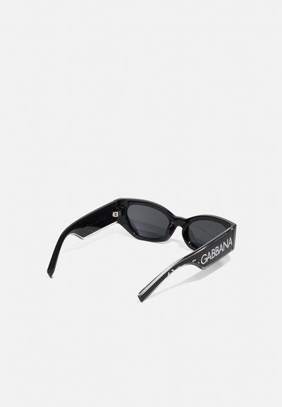 Óculos de sol negros com um design elegante e angular. Apresentam lentes escuras e uma marcação branca em destaque nas hastes. Material de plástico brilhante.