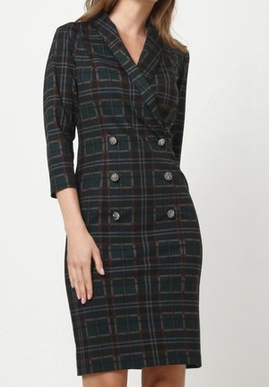 Mujer vestida con un vestido oscuro de cuadros con doble botonadura, seis botones decorativos y mangas tres cuartos, de pie contra un fondo liso.