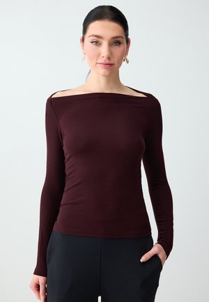 Donna con capelli scuri che indossa una maglia bordeaux a maniche lunghe con spalle scoperte e pantaloni neri, in piedi con una mano nella tasca davanti a uno sfondo semplice.