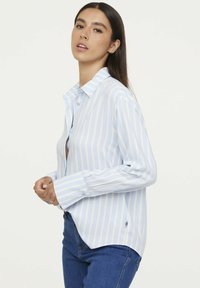 Light blue and white striped button-up shirt, long sleeves, relaxed fit, featuring a pointed collar and single logo detail at the hem.