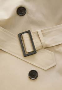 Beige trenchcoat stof met een gladde textuur, voorzien van een zwarte knop en een rechthoekige riemgesp met een metalen afwerking.
