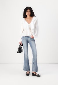 Blusa bianca con volant e laccio frontale, jeans azzurri a zampa d'elefante, borsa nera e ballerine nere con fori circolari.