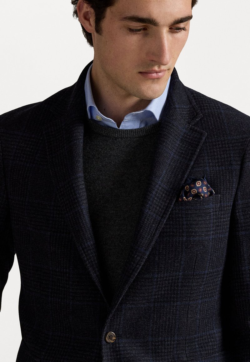 Blazer a quadri navy in tessuto texturizzato, camicia colletto azzurro chiaro e un maglione scuro sotto. Include un fazzoletto da taschino a fantasia.