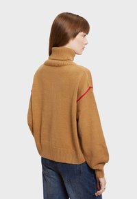 Beige gebreide turtleneck trui met een lose pasvorm, voorzien van een rode streep langs de schouder en geribde accenten op de manchetten en de zoom.