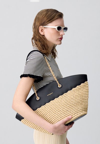 Borsa a tote di grandi dimensioni con una base in paglia testurizzata, parte superiore in pelle nera e bordo ondulato. Le cinghie a catena dorata aggiungono dettagli e contrasto.