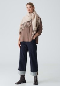Donna che indossa un sciarpa beige con frange sopra una camicia marrone larga con bottoni, jeans scuri a gamba larga con risvolti, e stivaletti neri, in piedi con una mano in tasca.