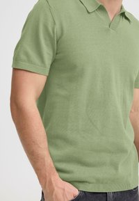 Polo manches courtes vert clair avec col en V, tissu en maille et coupe décontractée, associé à un jean foncé.